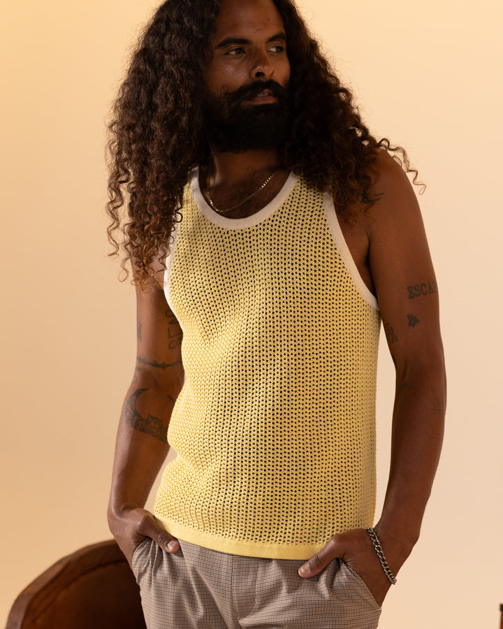 A bearded individual with long curly hair effortlessly styled in The Dominica Crochet Tank - Plantain by Dandy Del Mar and light brown pants stands with hands in pockets, exuding a relaxed fit.