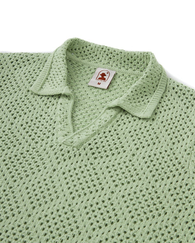 The Antibes Crochet Polo Shirt in Pistachio by Dandy Del Mar features a light green crochet knit, short sleeves, a textured diamond pattern, and a folded collar. Size "M" is tagged inside the neckline.