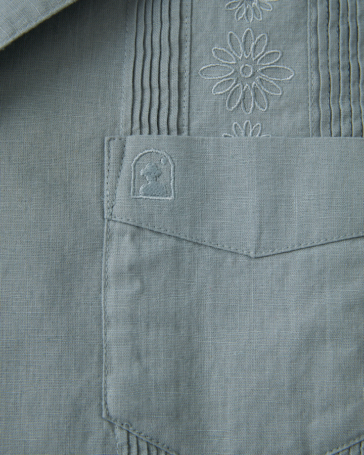 The pocket of a grey Dandy Del Mar Brisa Linen Shirt with embroidered flowers.