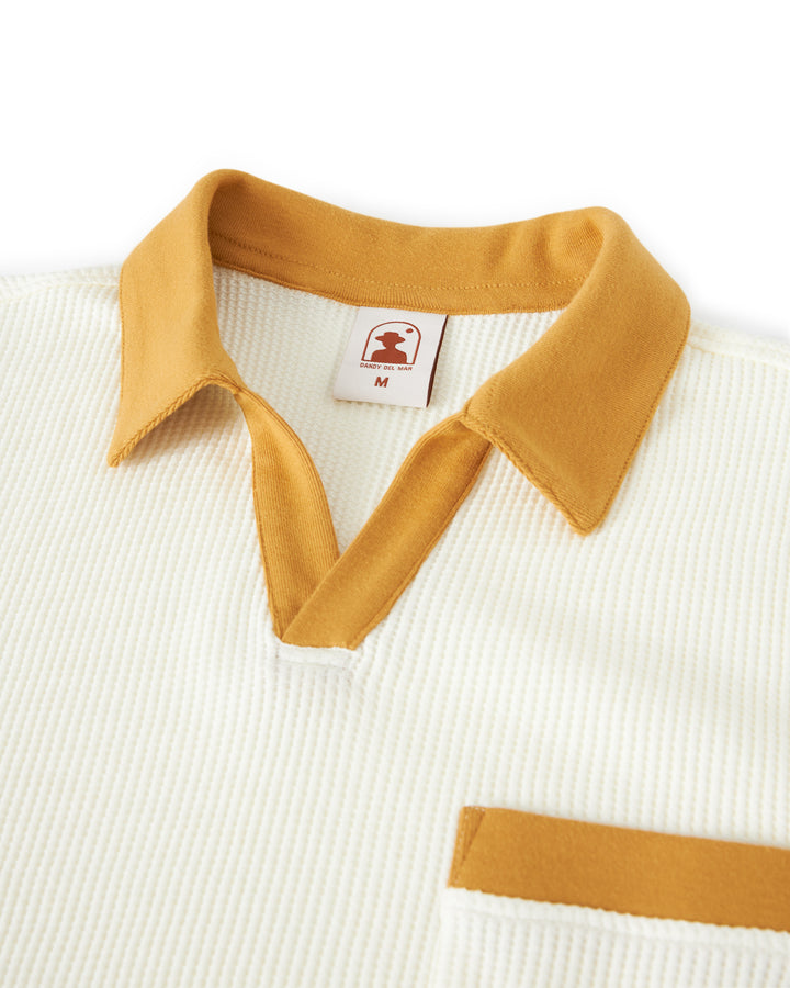 Close-up of a cream-colored The Cannes Waffle Knit Shirt - Vintage Ivory with a mustard buttonless polo collar and pocket trim, displaying a tag with the Dandy Del Mar logo and "M" size. The shirt features textured waffle knit fabric.