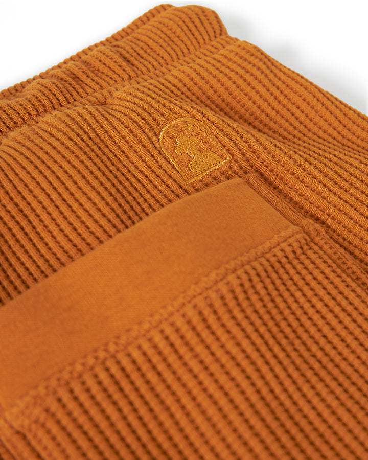 Close-up of a pair of rust-colored waffle knit fabric pants, featuring an embroidered logo above the back pocket and an elastic waistband. Dandy Del Mar's The Cannes Waffle Knit Shorts - Burnt Sienna.