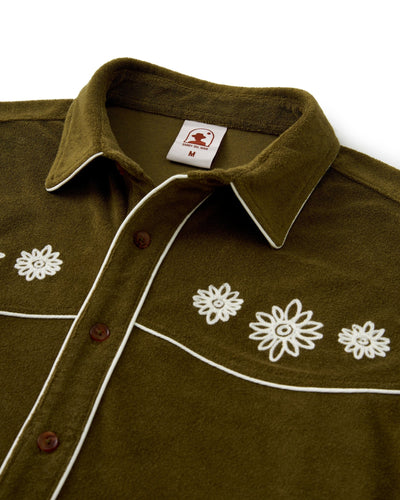 The Gaucho Terry Cloth Shirt - Arbequina by Dandy Del Mar features olive green terry cloth, white piping, brown buttons, and white floral chest embroidery. Size “M” label shows inside the collar.
