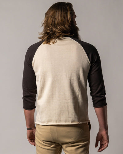 A man with long hair and a beard stands with his back to the camera, wearing the Dandy Del Mar Grenada Knit Baseball Tee - Tierra in cream with dark sleeves, paired with tan pants.