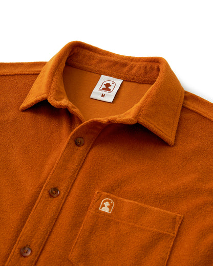 The Tropez Shirt - Burnt Sienna by Dandy Del Mar is made of terry cloth and includes an orange collar, brown buttons, a chest pocket with a silhouette logo, and another logo inside the collar. Ideal for sunny Côte d'Azur days, it is shown against a white background.