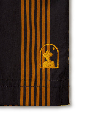 Close-up of the hem from Dandy Del Mar’s The Ventura Volley Shorts - Albatross, featuring vertical brown stripes on black and a yellow embroidered logo with a silhouette inside an arch and a small dot above.