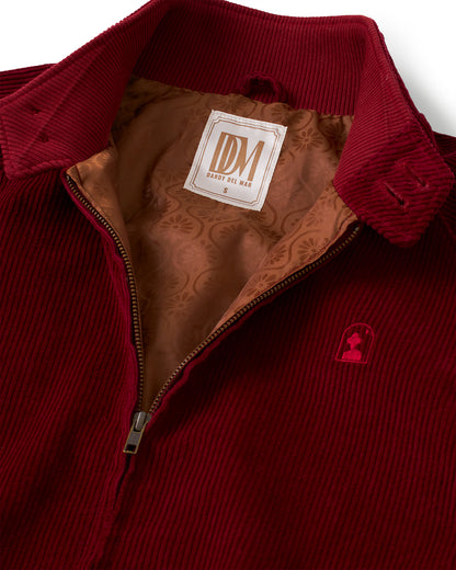 Close-up of the Corsica Corduroy Jacket by Dandy Del Mar in Sangria, featuring a zip, a beige patterned lining reminiscent of Gardenia jacquard silk, and a visible label reading "DDM," along with a small embroidered red emblem on the chest.