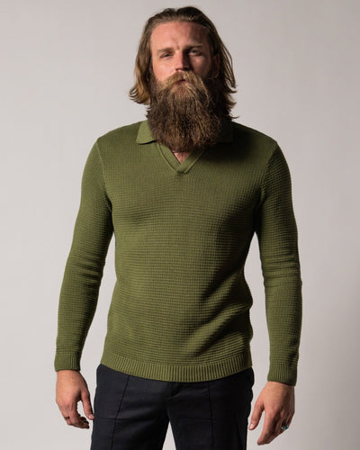A man with long hair and a full beard wears the Dandy Del Mar Sebastian LS Polo Shirt in Arbequina, paired with dark pants, standing before a plain light background.