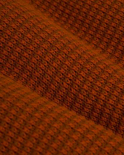 Close-up of The Sebastian Long Sleeve Polo in Burnt Sienna by Dandy Del Mar, showcasing a brown textured knit fabric with a repeating wavy pattern, reminiscent of an intricate design.