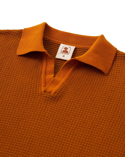 A close-up of The Sebastian LS Polo Shirt in Burnt Sienna by Dandy Del Mar, a men’s textured knit sweater polo with a collar, V-neck opening, and visible size and brand label.