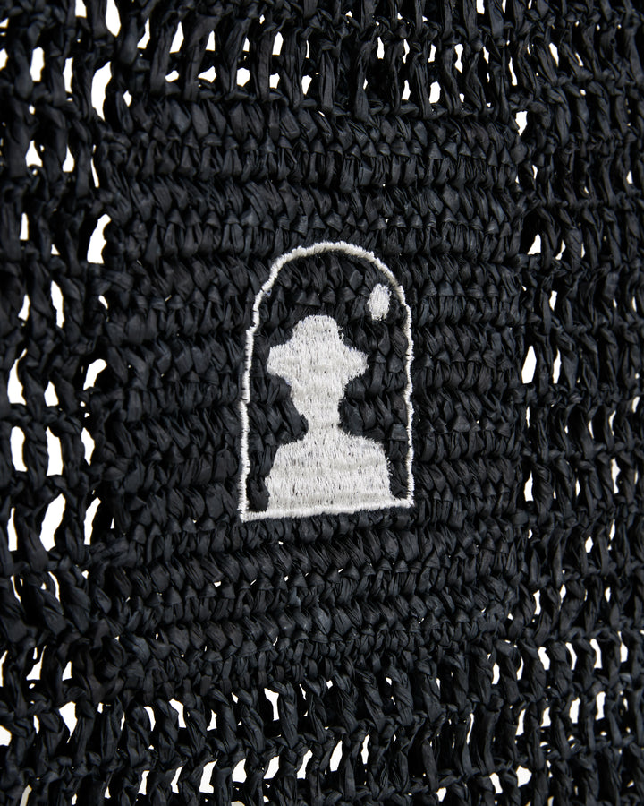 Close-up of a black knitted fabric with a white and gray embroidered Dandy Del Mar logo, featuring stylized animal silhouette in raffia-effect yarn.
