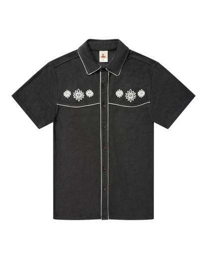 The Gaucho Shirt - Onyx by Dandy Del Mar, a short-sleeved black terry cloth shirt with white piping and embroidered floral chest, is shown laid flat on a black background.