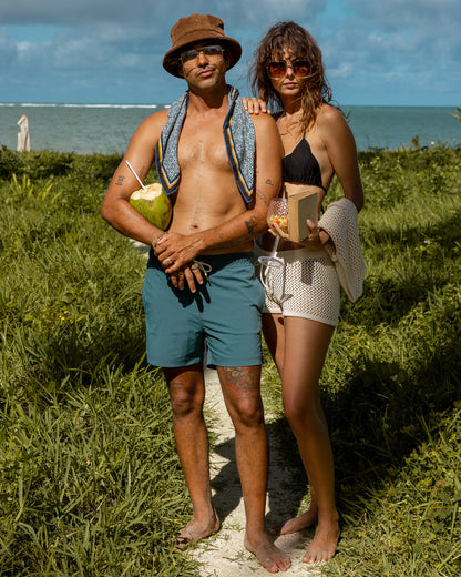 A man and woman stand on a grassy seaside path. The man, wearing The Ventura Volley Short - Acqua by Dandy Del Mar, a hat, and sunglasses, holds a coconut. The woman dons a nylon spandex bikini top and shorts while holding an apple and book.