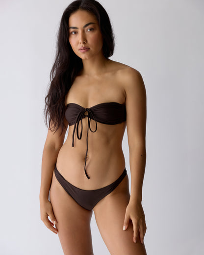 A woman stands against a plain background wearing the Dandy Del Mar Biarritz Top in Tierra, a dark brown tie front bandeau bikini with chic cinch detailing at the front.