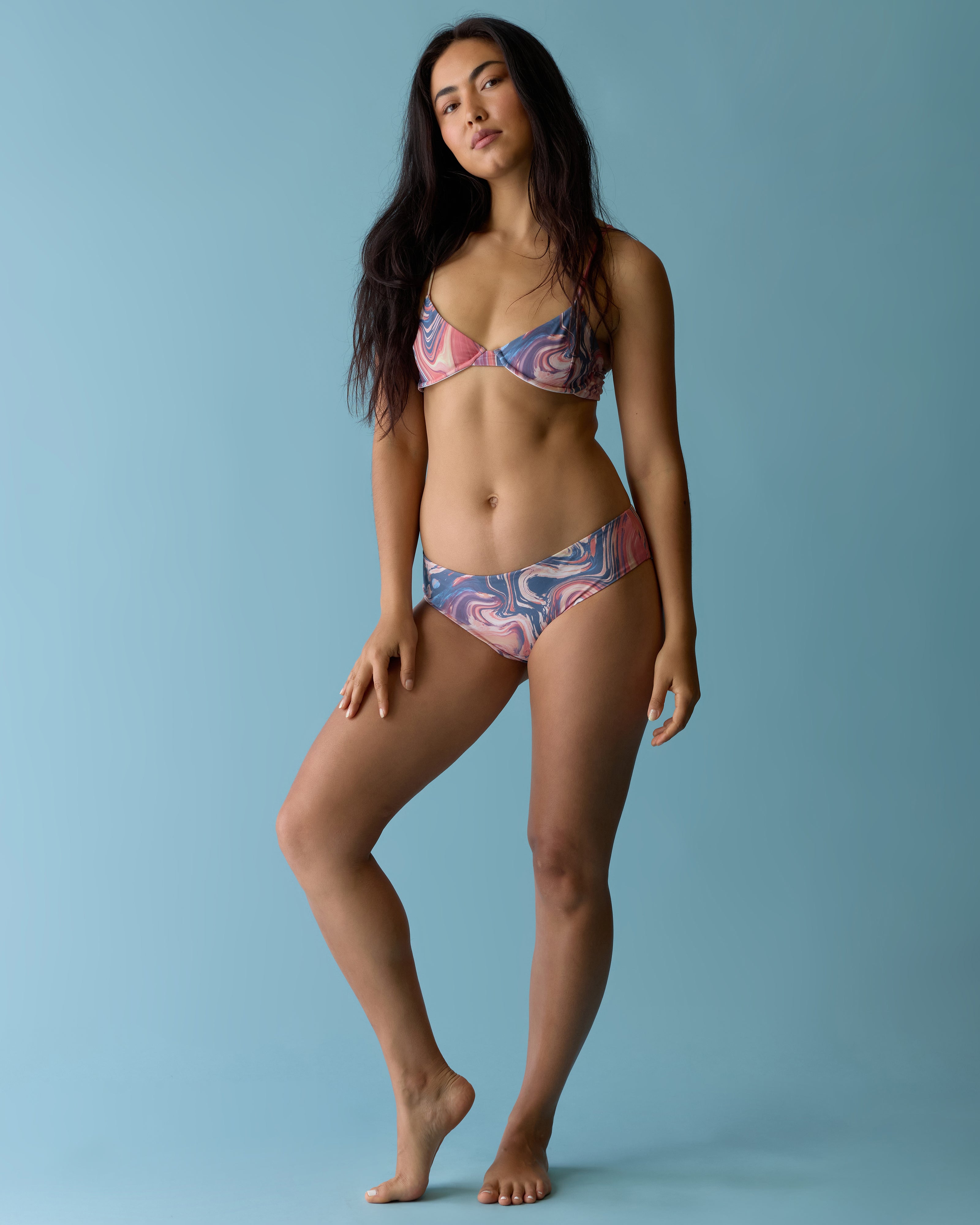 A woman stands on one leg, modeling The Delphi Bottoms - Lantana by Dandy Del Mar, in a vibrant marbled print, set against a plain light blue background.