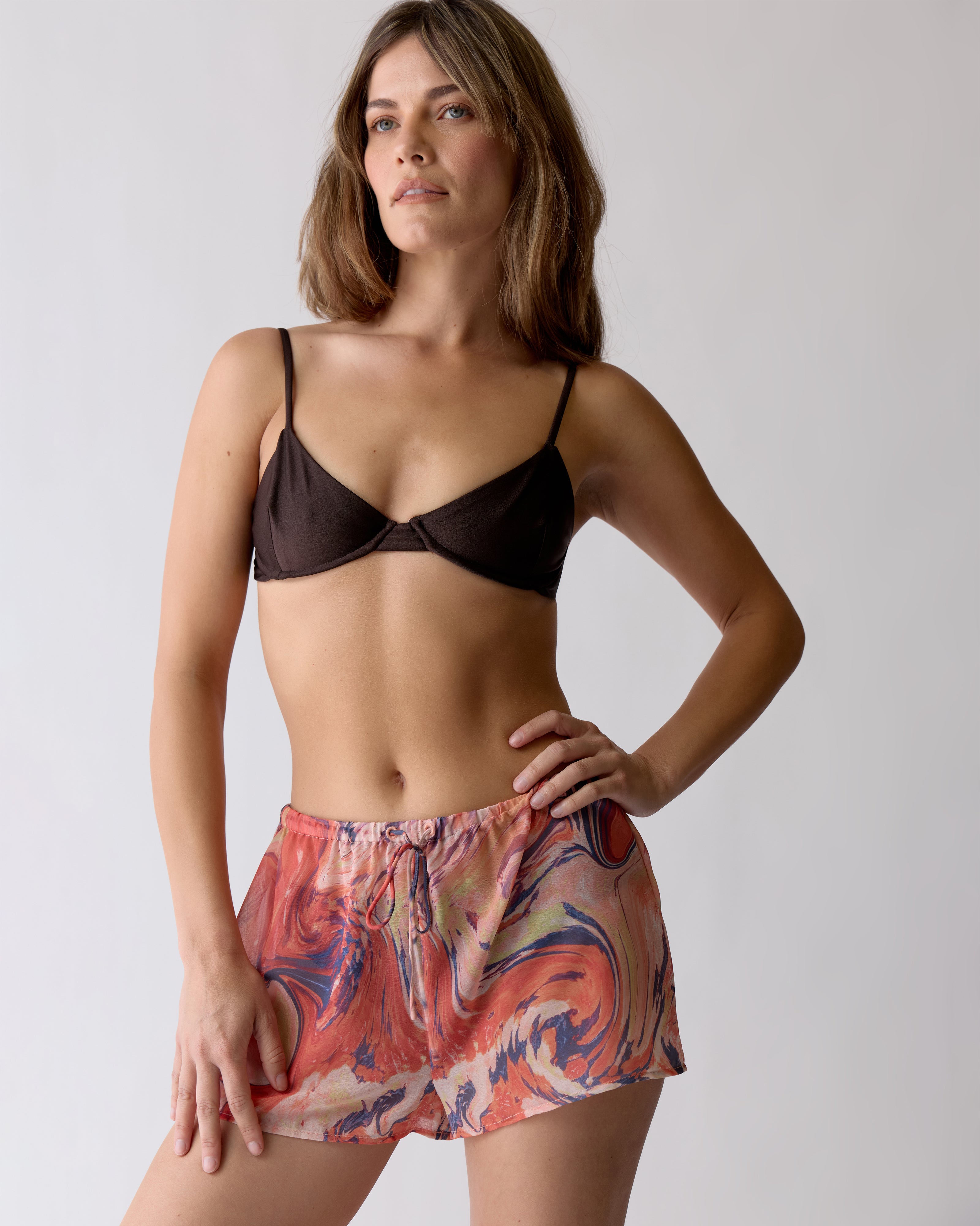 A woman stands against a plain background wearing the Dandy Del Mar Avila Top - Tierra—a dark brown bralette made from recycled nylon—paired with colorful, marbled shorts.