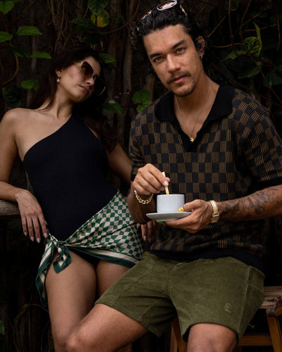 A man relaxes outdoors in the Dandy Del Mar Sebastian Knit Polo Shirt in Onyx, holding a cup and saucer, while a woman in sunglasses and a one-shoulder swimsuit leans against a chair beside him.