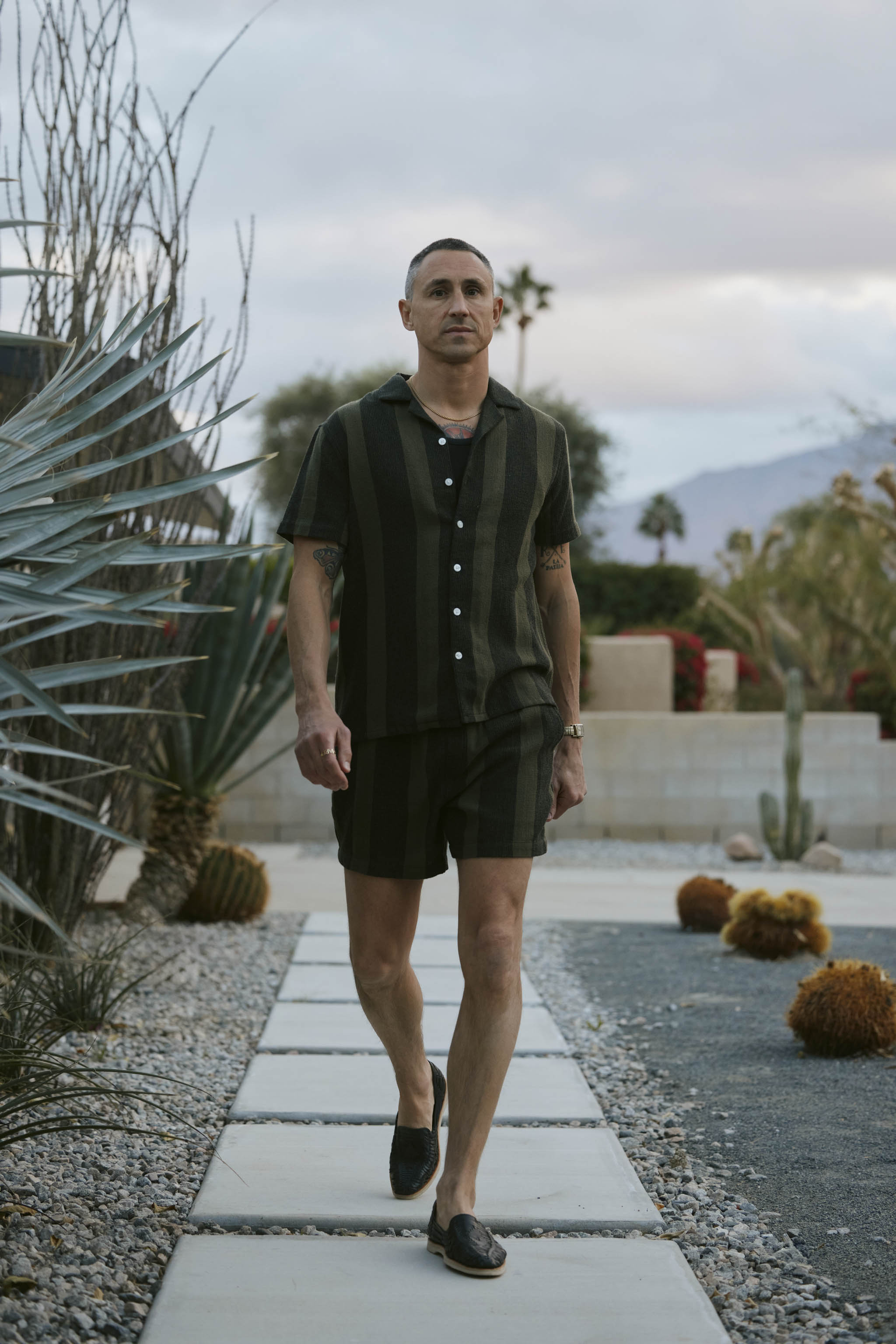 A person strolls along a stone path in a desert garden, sporting The Palma Shirt in Onyx by Dandy Del Mar, accompanied by shorts. Behind them are desert flora and a cloudy sky.