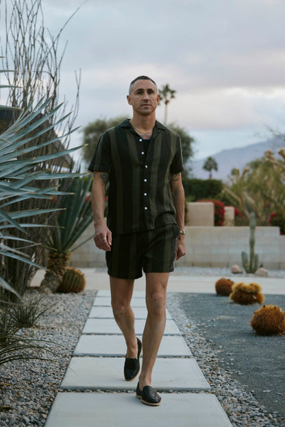 A person strolls along a stone path in a desert garden, sporting The Palma Shirt in Onyx by Dandy Del Mar, accompanied by shorts. Behind them are desert flora and a cloudy sky.