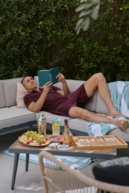 A man, wearing The Rio Velour Short in Bordeaux from Dandy Del Mar, relaxes on a beige outdoor sofa while reading a book. Nearby, a table holds snacks, drinks, and a backgammon set against a backdrop of lush green foliage.
