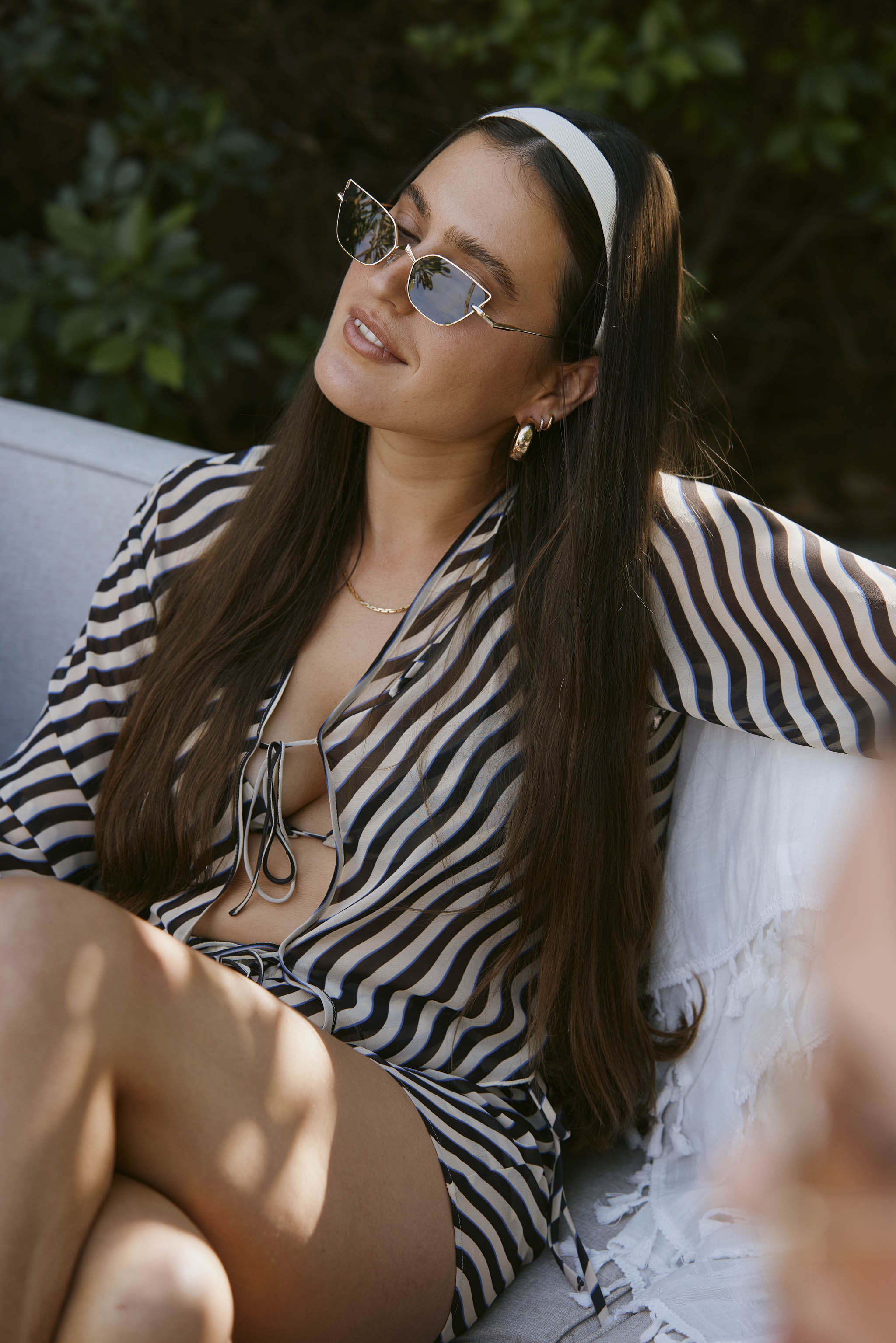 A woman with long hair in sunglasses and a striped Curacao Top - Tierra by Dandy Del Mar sits on an outdoor sofa, smiling with one arm resting on the back.
