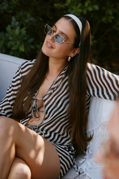 A woman with long hair in sunglasses and a striped Curacao Top - Tierra by Dandy Del Mar sits on an outdoor sofa, smiling with one arm resting on the back.