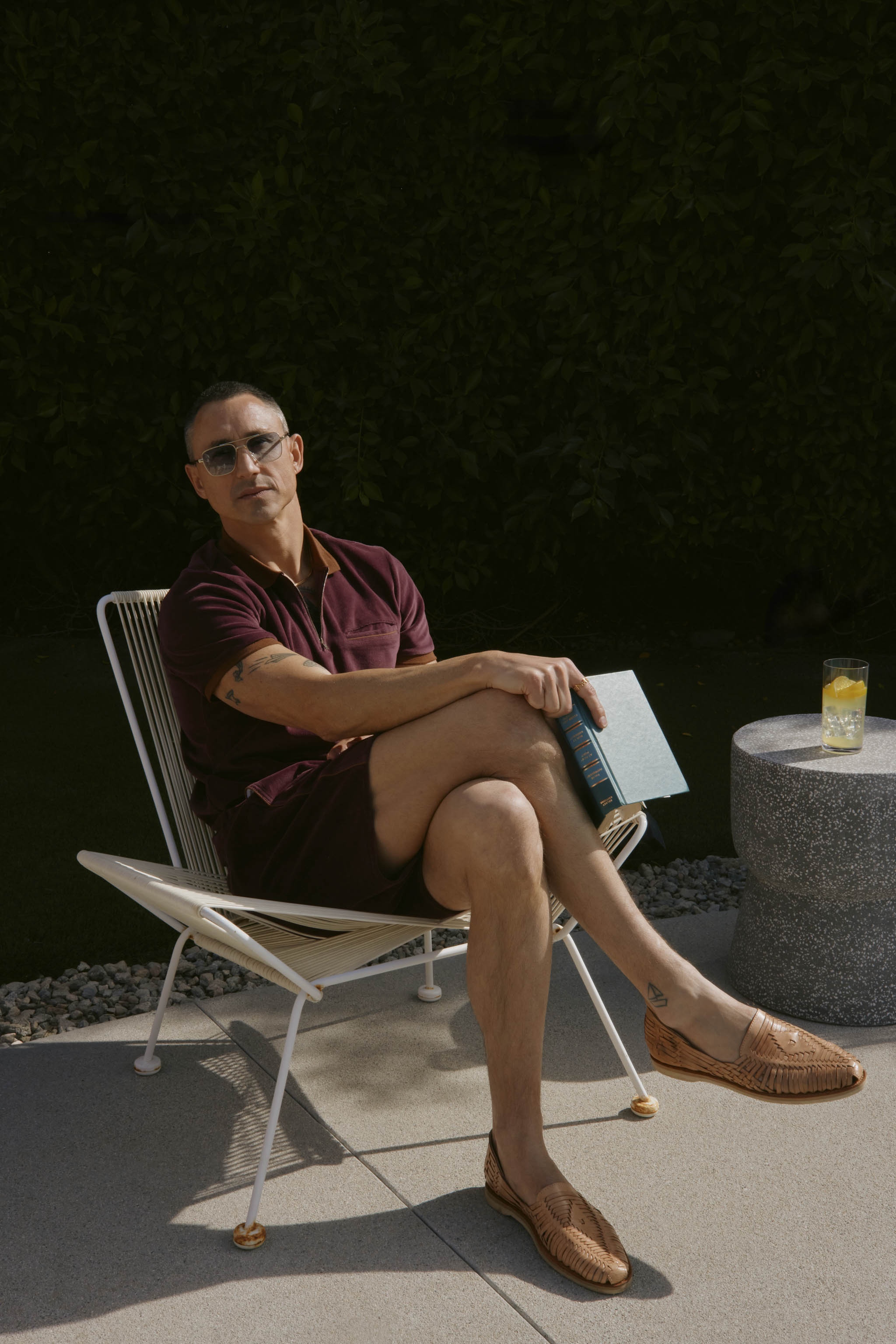 A person in sunglasses sits outdoors on a wire chair, holding a book. They wear The Rio Velour Short in Bordeaux by Dandy Del Mar and brown woven shoes. A drink is placed on a small circular table nearby.
