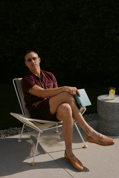 A person in sunglasses sits outdoors on a wire chair, holding a book. They wear The Rio Velour Short in Bordeaux by Dandy Del Mar and brown woven shoes. A drink is placed on a small circular table nearby.