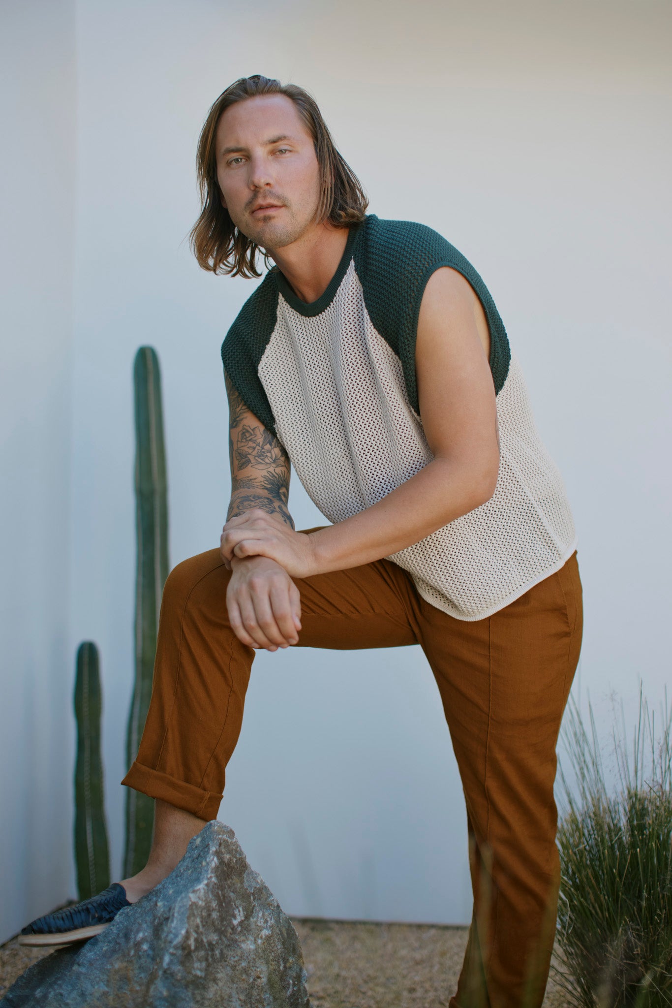 A man with shoulder-length hair poses outdoors, one leg on a rock, wearing Dandy Del Mar’s The Porto Crochet Tank in Sabbia with brown pants. Cacti and grass appear in the background.