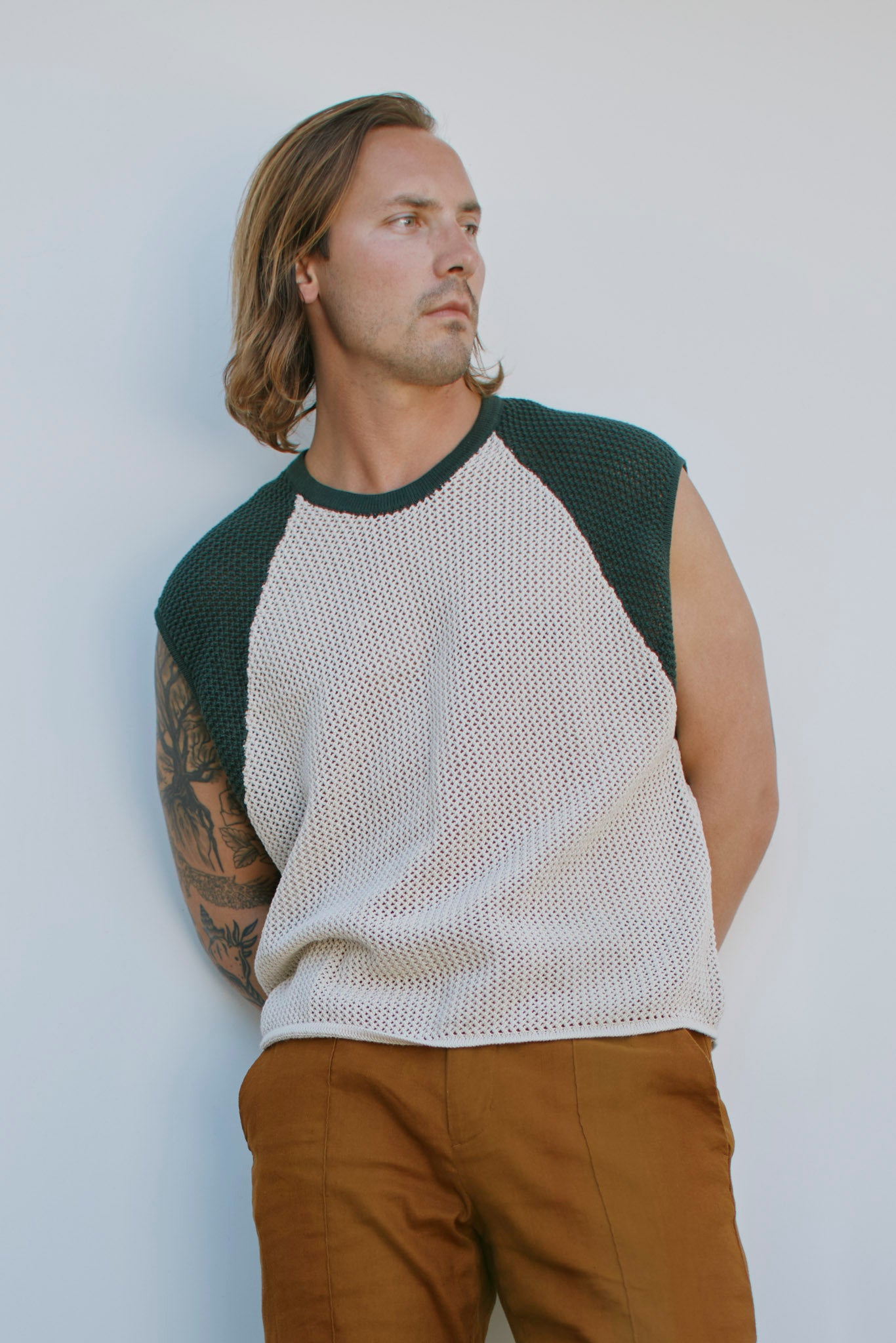 A man with shoulder-length hair and an arm tattoo wears The Porto Crochet Tank in Sabbia by Dandy Del Mar, styled with dark green sleeves and brown pants, posing against a plain light background.