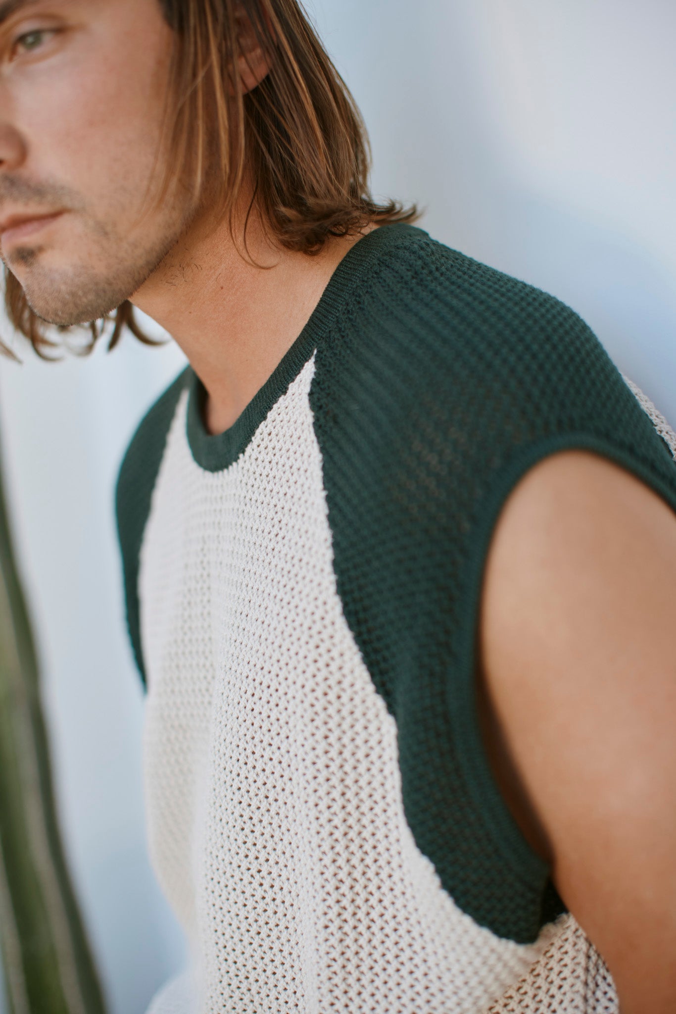 A person with shoulder-length brown hair wears The Porto Crochet Tank - Sabbia by Dandy Del Mar, a sleeveless cropped top with dark green trim and white mesh fabric, against a softly blurred background.