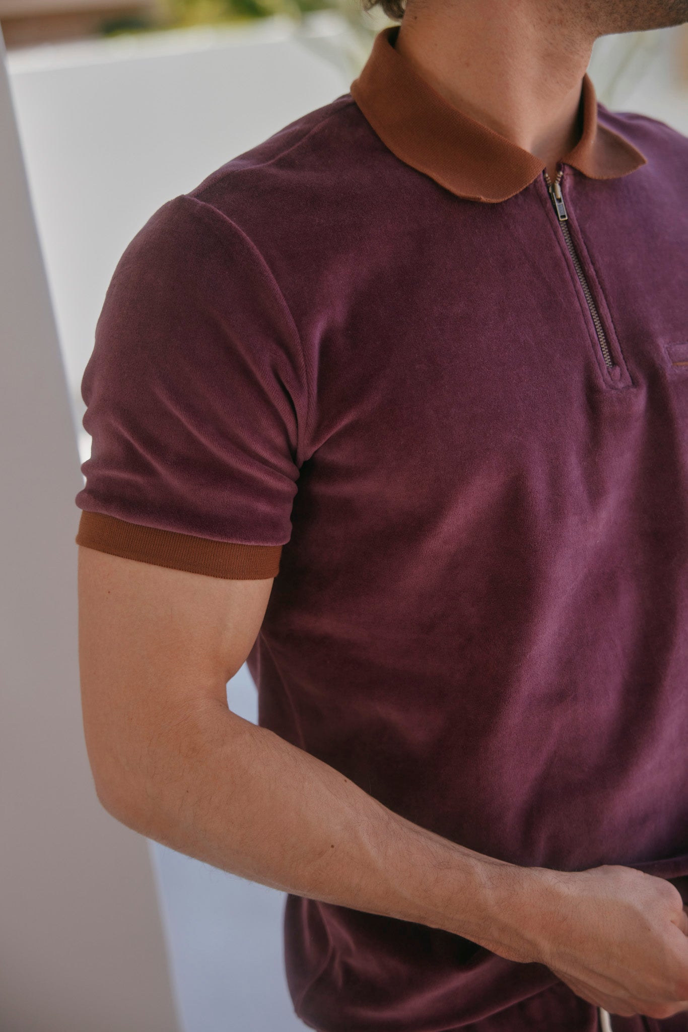 A man wears The Rio Velour Shirt - Bordeaux by Dandy Del Mar, a short-sleeve maroon polo with a brown collar and zipper, shown from shoulders to mid-torso.