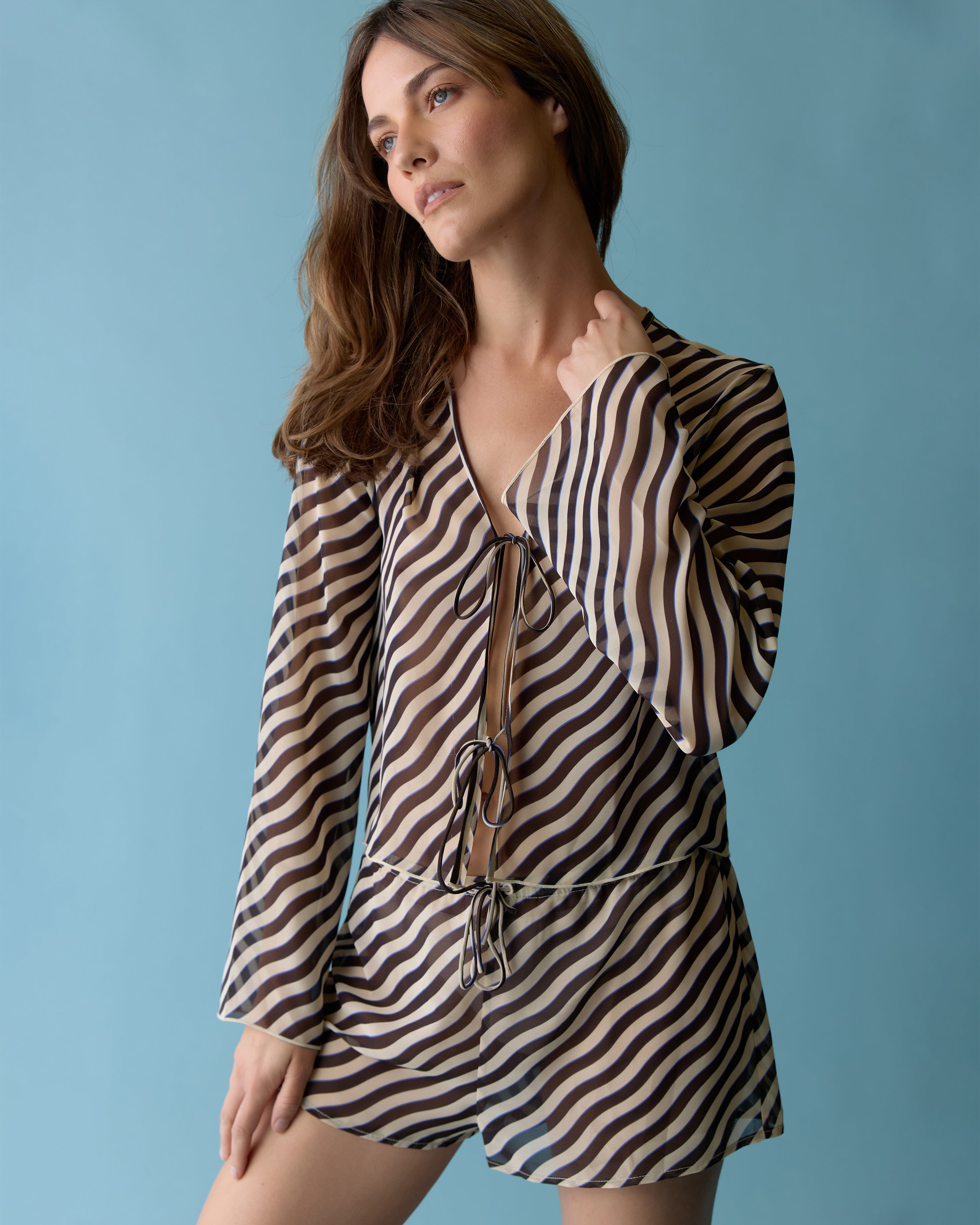 A woman wears The Curacao Top - Tierra by Dandy Del Mar, featuring long sleeves and a V-neck with an exotic black and beige wavy stripe print, standing against a blue background.