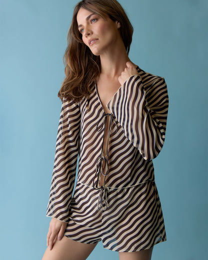 A woman wears The Curacao Top - Tierra by Dandy Del Mar, featuring long sleeves and a V-neck with an exotic black and beige wavy stripe print, standing against a blue background.