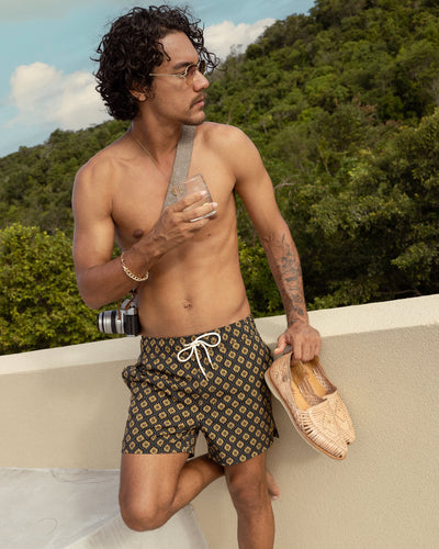 A shirtless man in Dandy Del Mar's The Ventura Volley Shorts—Oro Gardenia Print, with an elastic waist, stands by a low wall holding a drink and camera, wearing sunglasses and woven shoes, with greenery in the background.