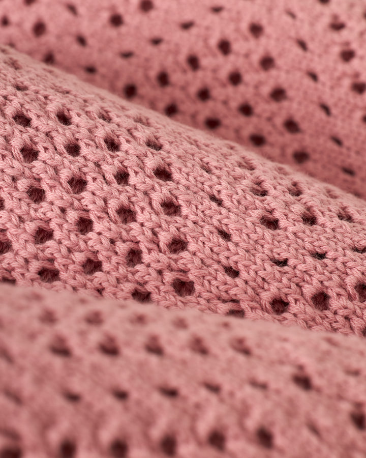 Close-up of The Antibes Crochet Shirt - Dawn by Dandy Del Mar, with pink knitted fabric featuring a pattern of small holes for a textured, airy appearance.