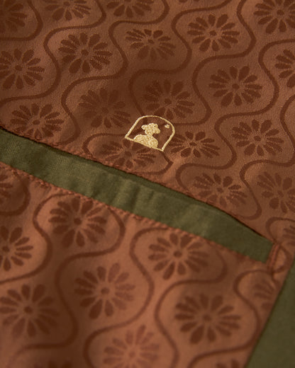 A close-up of Dandy Del Mar's The Brisa Linen Blazer - Berdea showcases its brown fabric adorned with floral patterns and a stitched green strip, featuring a small golden silhouette logo.