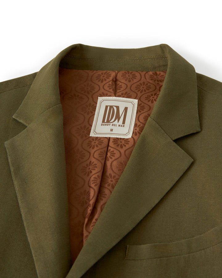 Close-up of an olive-green linen blazer with a brown jacquard-patterned inner lining. The label reads "Dandy Del Mar The Brisa Linen Blazer - Berdea.