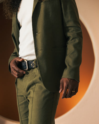 Person wearing an olive green suit with a white shirt, showcasing a studded belt and a ring on their left hand. The Brisa Linen Trouser - Berdea by Dandy Del Mar offers a tailored fit, seamlessly blending style and comfort.