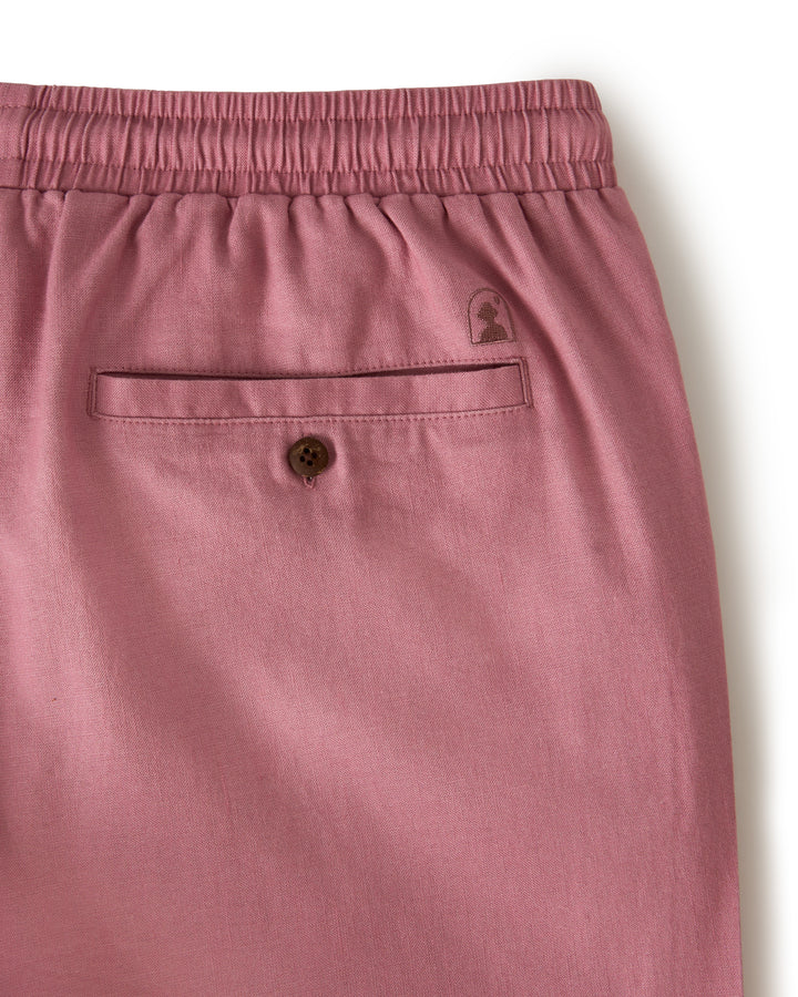 Close-up of The Brisa Linen Pant - Dawn by Dandy Del Mar in pink, showcasing an elastic waistband, buttoned back pocket, and a subtle logo above the pocket—ideal for comfortable vacation wear.