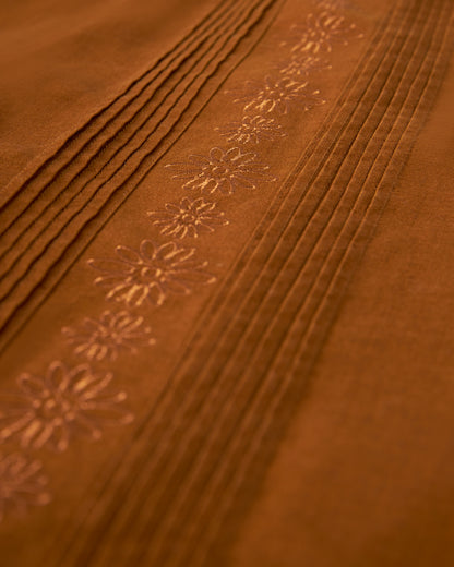 Close-up of an orange-brown Brisa Linen Shirt - Sedona by Dandy Del Mar, featuring gardenia floral embroidery and several parallel pleats, accented by coconut shell buttons.