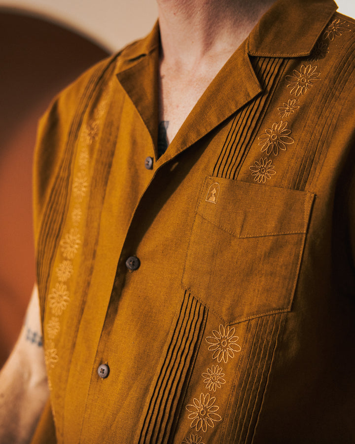 Close-up of a person wearing The Brisa Linen Shirt - Sedona by Dandy Del Mar, showcasing its brown embroidered guayabera style with gardenia floral patterns and pleats. This shirt features a pocket and coconut shell buttons, adding a touch of rustic charm.