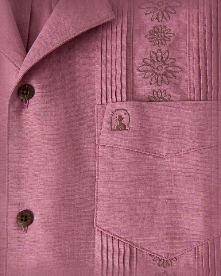 Close-up of The Brisa Linen Shirt - Dawn by Dandy Del Mar, showcasing brown floral embroidery on a pink fabric and a pocket with a silhouette design.