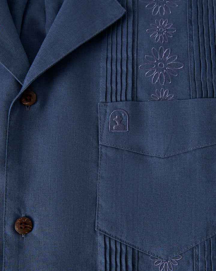 A close-up of The Brisa Linen Shirt - Moontide by Dandy Del Mar, featuring blue fabric with floral embroidery and vertical pleats. It includes brown buttons, a front pocket, and a small embroidered logo.