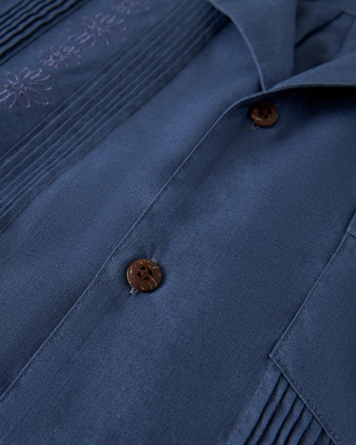 A close-up of The Brisa Linen Shirt - Moontide by Dandy Del Mar shows brown buttons and floral embroidery near the collar.