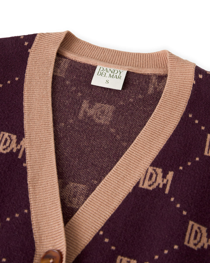 Close-up of The Canela Cardigan - Amethyst, featuring a rich dark purple cotton design with tan trim, highlighting the "Dandy Del Mar" label and an eye-catching monogram pattern with letters.