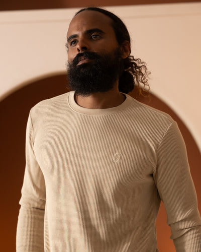 A bearded man with tied-back hair wears the Dandy Del Mar The Cannes LS Waffle Knit Shirt in Truffle, standing indoors before an arched backdrop.