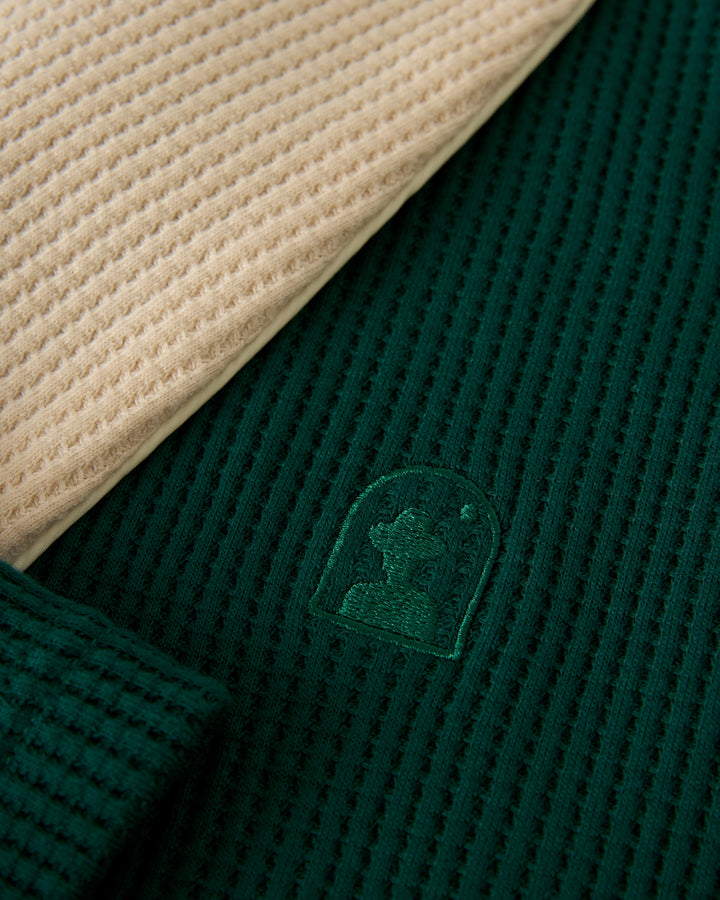 Close-up of two waffle-knit fabrics, one beige and the other from Dandy Del Mar's The Cannes Waffle Knit Robe in Albero green, featuring an embroidered arch-shaped logo.