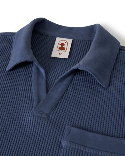 A close-up of the Dandy Del Mar Cannes Waffle Knit Polo Shirt in Moontide, showcasing its waffle knit fabric, chest pocket, and an inner collar tag with a brown logo and size "M".