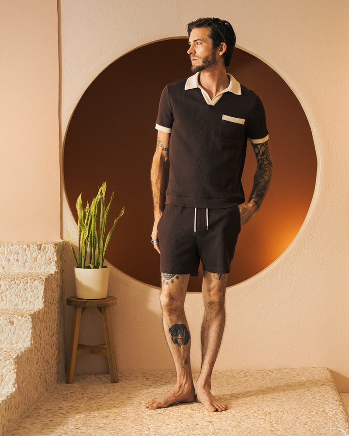 A barefoot man with tattoos stands on a step in front of a round wall feature, wearing a brown short-sleeve waffle knit shirt and The Cannes Waffle Knit Shorts in Carajillo from Dandy Del Mar, which feature an elastic waistband. A potted plant sits on a stool beside him.