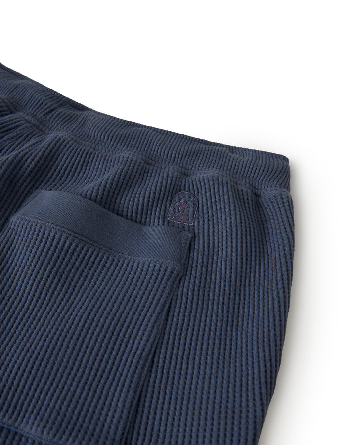 Close-up of Dandy Del Mar's The Cannes Waffle Knit Shorts in Moontide, featuring a dark blue textured fabric with a pocket and small emblem.